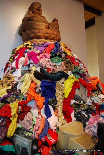Buddha sitting atop a pile of clothes and other junk.