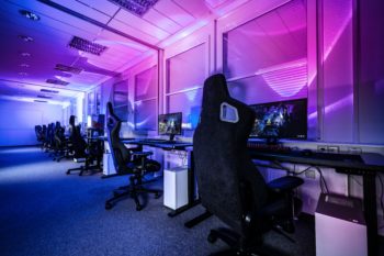 An empty room filled with computers and gamer chairs. The room is lit with blue and pink lighting creating a mood effect.