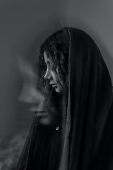 A black and white image of a person who appears female. They wear a long shawl over their head and gaze downward at the ground. They are in profile perspective facing to their right.